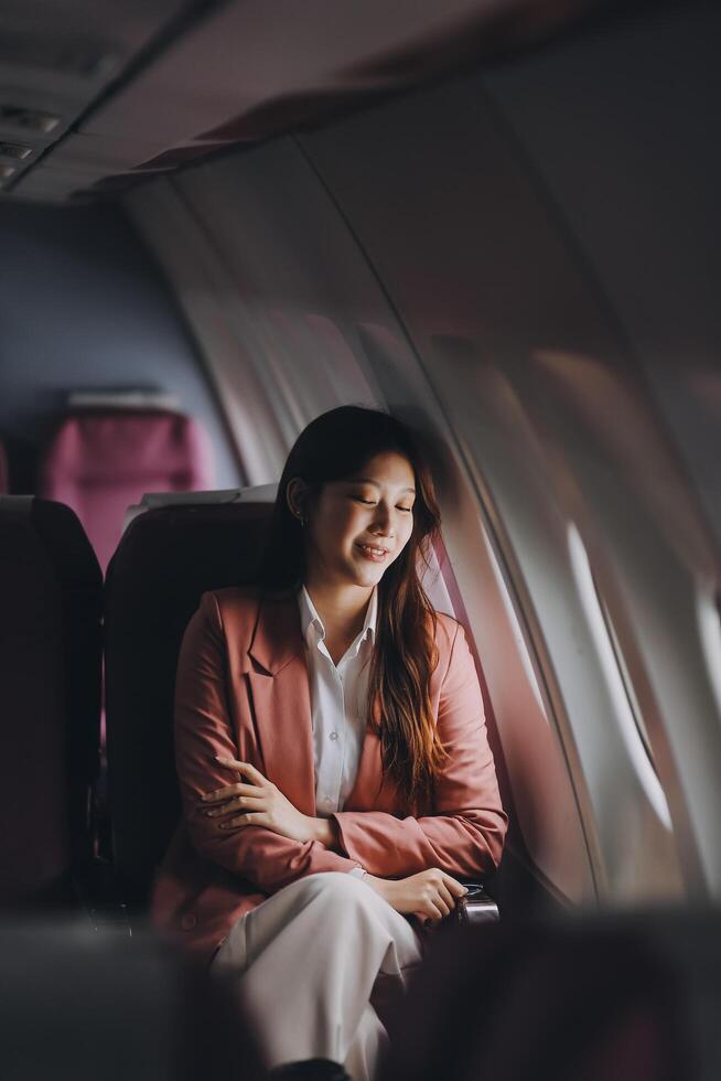 Portrait of a successful Asian businesswoman or entrepreneur in a formal suit on an airplane sitting in business class using a phone, computer laptop. Travel in style, work with grace. photo