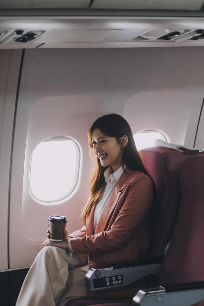 Portrait of a successful Asian businesswoman or entrepreneur in a formal suit on an airplane sitting in business class using a phone, computer laptop. Travel in style, work with grace. photo