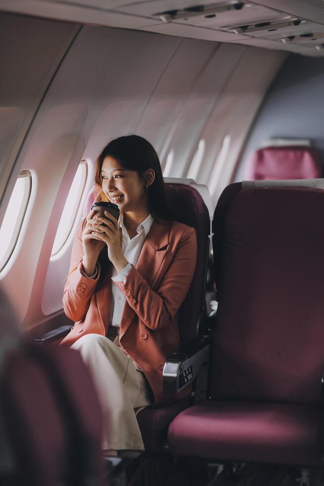 Portrait of a successful Asian businesswoman or entrepreneur in a formal suit on an airplane sitting in business class using a phone, computer laptop. Travel in style, work with grace. photo