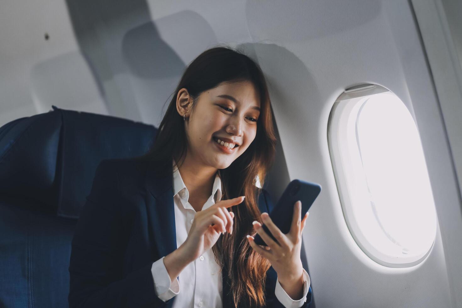 A young Asian female airplane passenger sits by the window during the flight, holding her boarding pass, with her carry-on luggage stored overhead, wearing her seatbelt, and enjoying the journey. photo