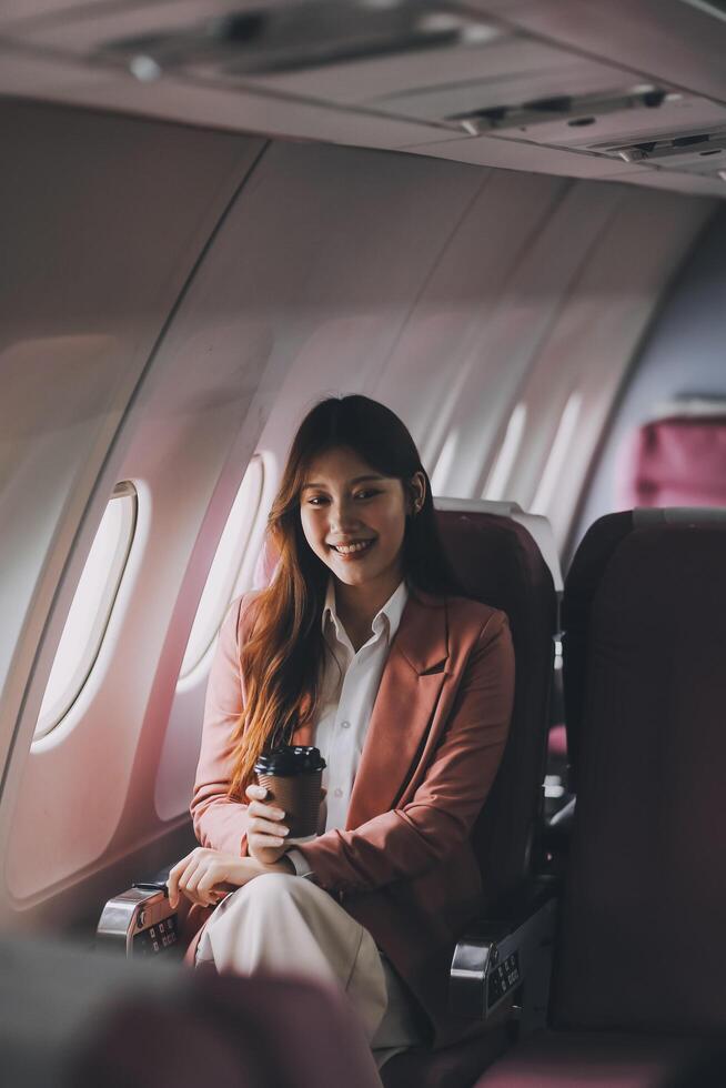 Portrait of a successful Asian businesswoman or entrepreneur in a formal suit on an airplane sitting in business class using a phone, computer laptop. Travel in style, work with grace. photo