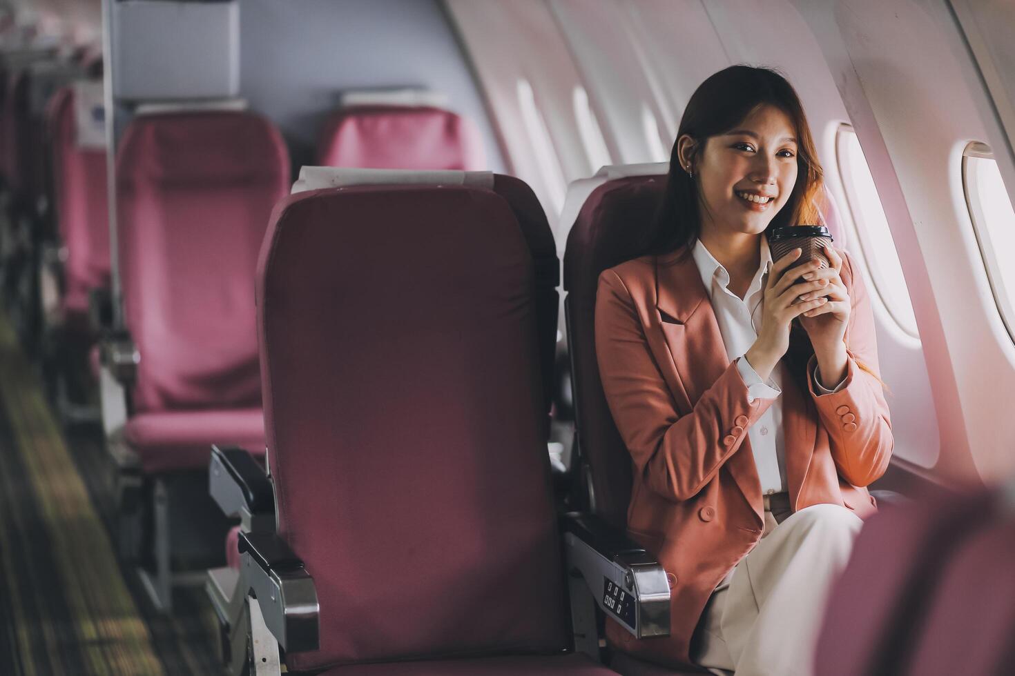 Portrait of a successful Asian businesswoman or entrepreneur in a formal suit on an airplane sitting in business class using a phone, computer laptop. Travel in style, work with grace. photo