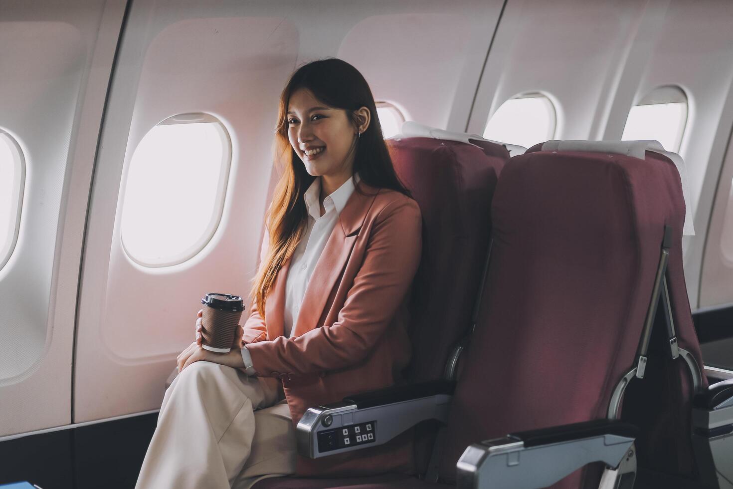 Portrait of a successful Asian businesswoman or entrepreneur in a formal suit on an airplane sitting in business class using a phone, computer laptop. Travel in style, work with grace. photo