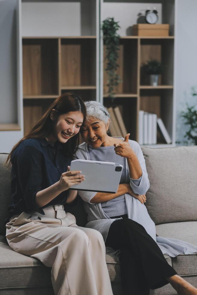 Happy grownup daughter showing content on tablet to mature mother. Two family generations women with digital computer gadget resting on couch together, watching videos, making call photo