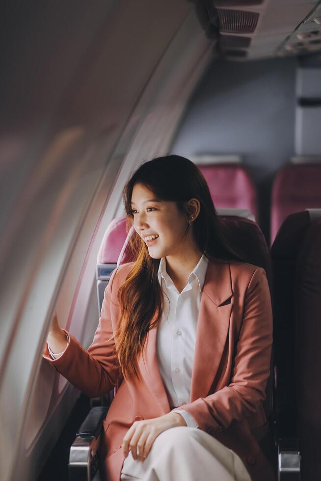 Portrait of a successful Asian businesswoman or entrepreneur in a formal suit on an airplane sitting in business class using a phone, computer laptop. Travel in style, work with grace. photo