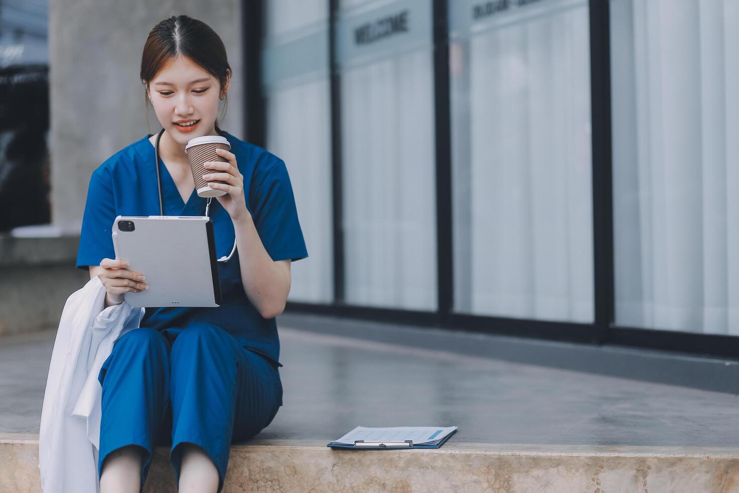 A cheerful woman in scrubs holds a clipboard, standing confidently before a modern hospital building, representing professional healthcare service. photo