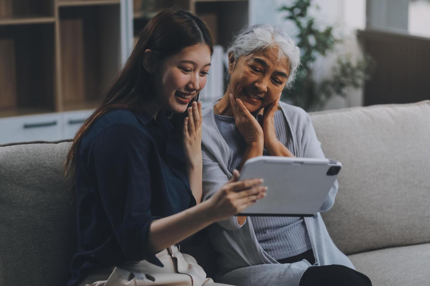 Happy grownup daughter showing content on tablet to mature mother. Two family generations women with digital computer gadget resting on couch together, watching videos, making call photo