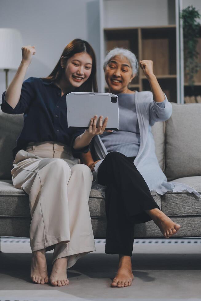 Happy grownup daughter showing content on tablet to mature mother. Two family generations women with digital computer gadget resting on couch together, watching videos, making call photo