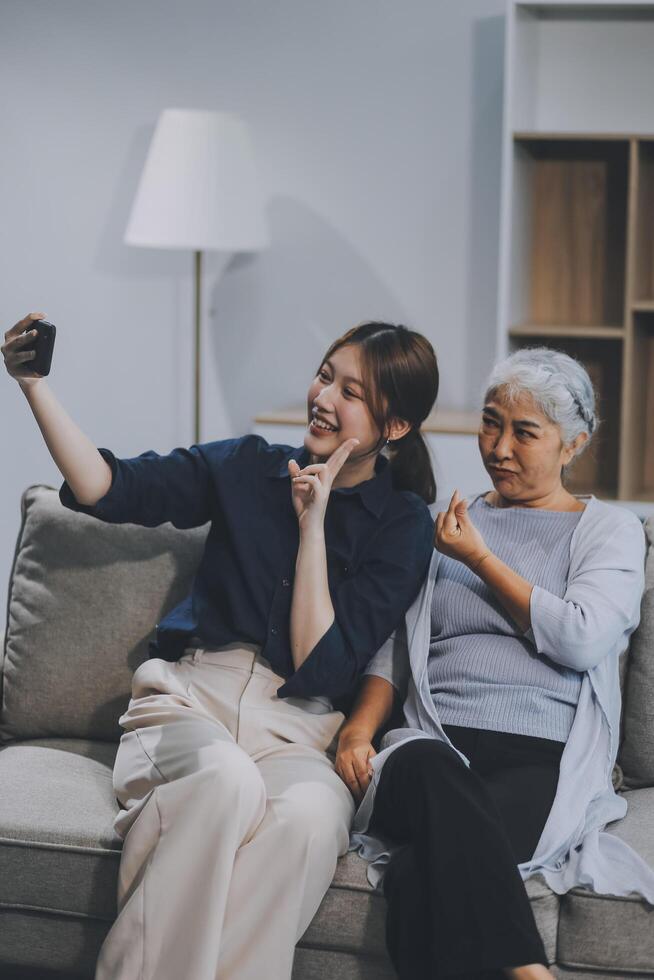 Cheerful mother and pretty teenager kid relaxing at cozy home, using mobile phone for Internet communication, resting on sofa with digital gadget, enjoying leisure, looking at camera photo