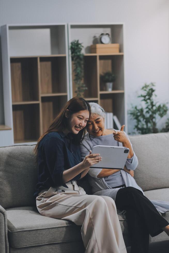 Happy grownup daughter showing content on tablet to mature mother. Two family generations women with digital computer gadget resting on couch together, watching videos, making call photo
