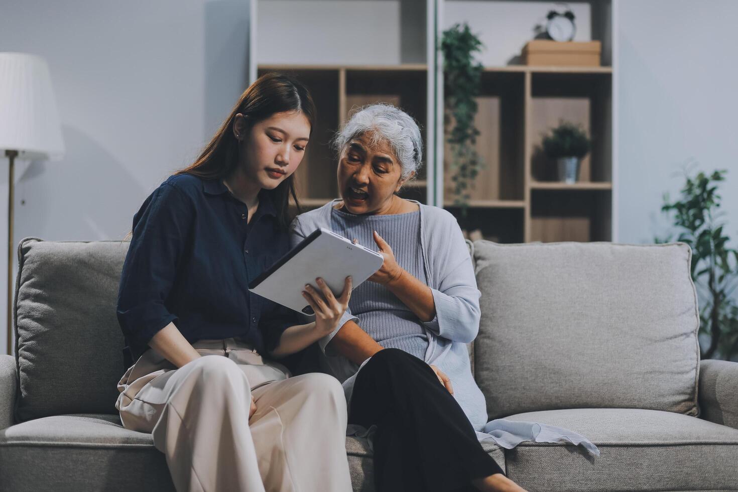 Happy grownup daughter showing content on tablet to mature mother. Two family generations women with digital computer gadget resting on couch together, watching videos, making call photo