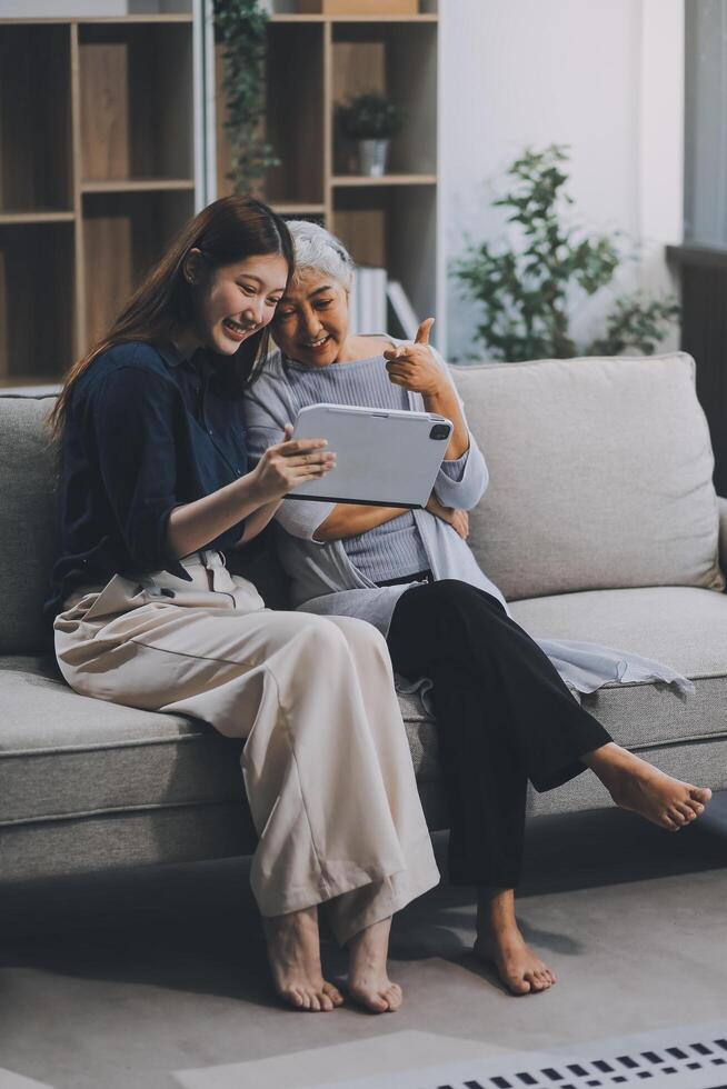 Happy grownup daughter showing content on tablet to mature mother. Two family generations women with digital computer gadget resting on couch together, watching videos, making call photo