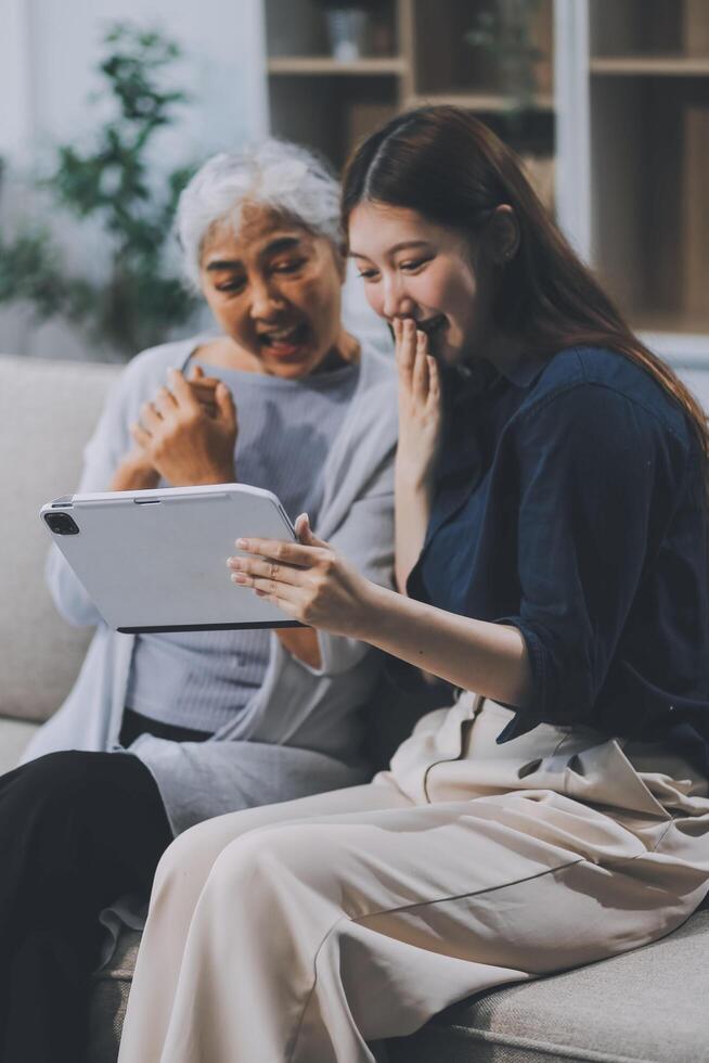 Happy grownup daughter showing content on tablet to mature mother. Two family generations women with digital computer gadget resting on couch together, watching videos, making call photo