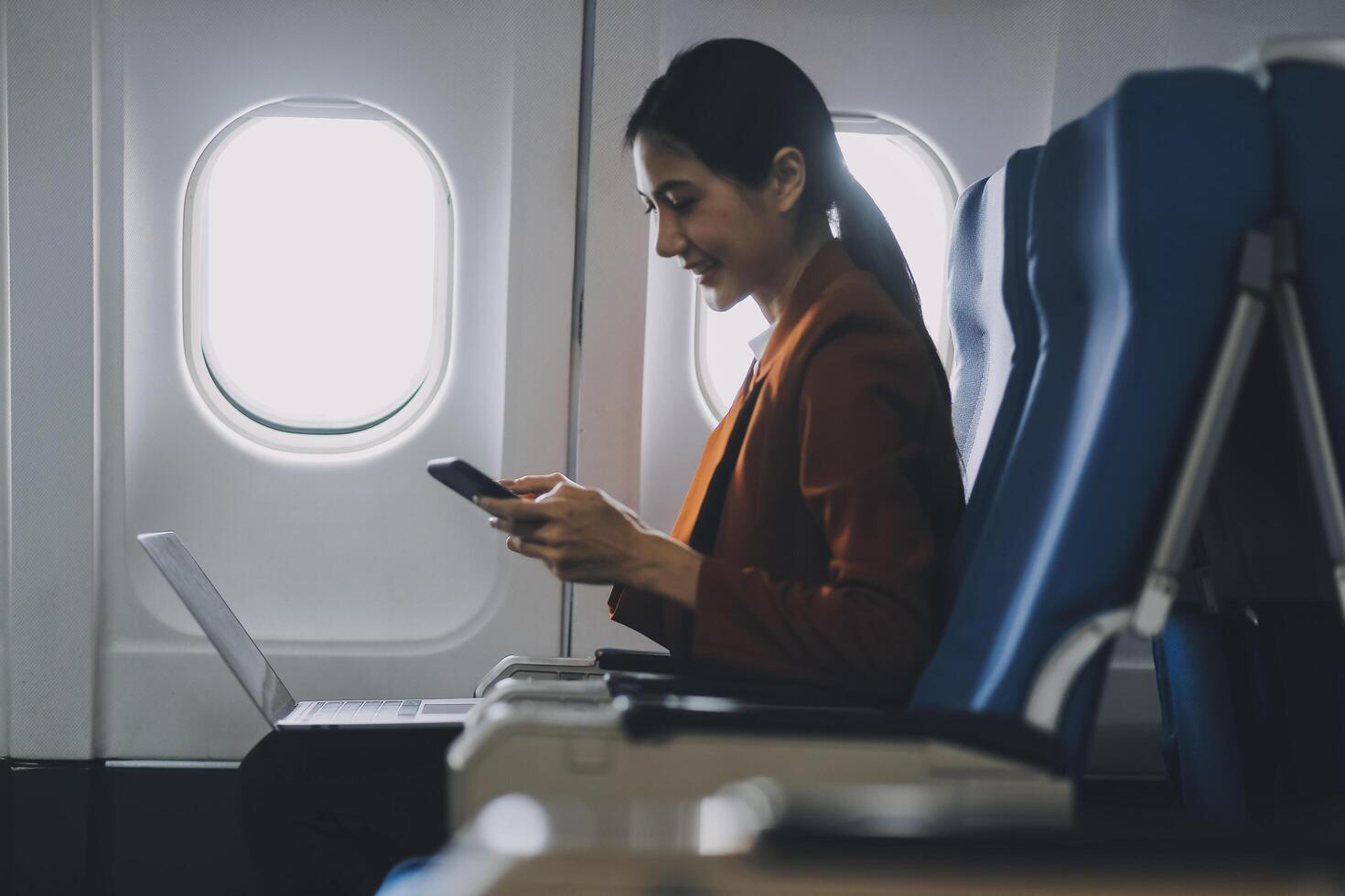 Asian female tourist checks notifications on smartphone while seated on airplane A businesswoman happily shares media from her phone, taking photos of the view from the window during a flight.