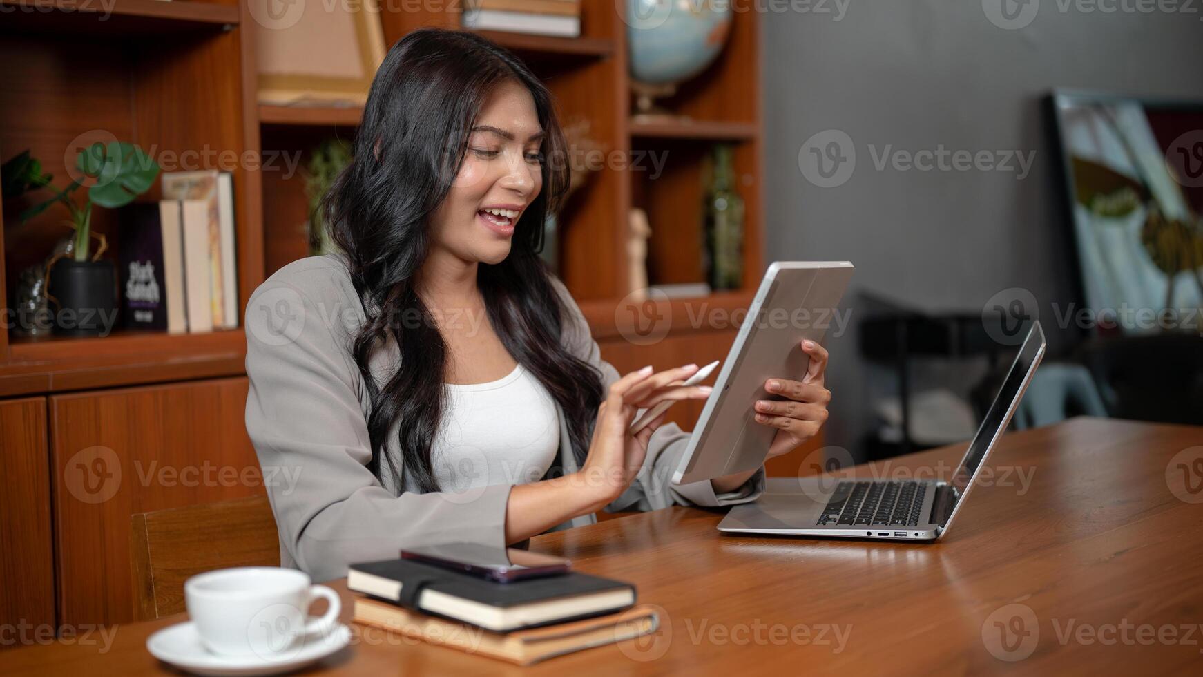 A positive Asian businesswoman using a digital tablet while sitting in front of a laptop in office. photo