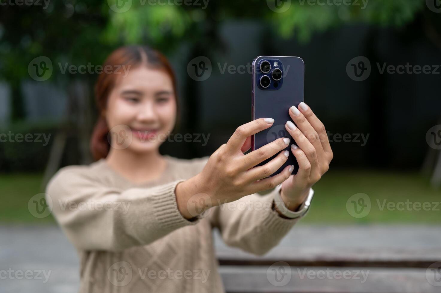 A positive Asian woman is taking a selfie or talking on a call on a bench in a park. photo