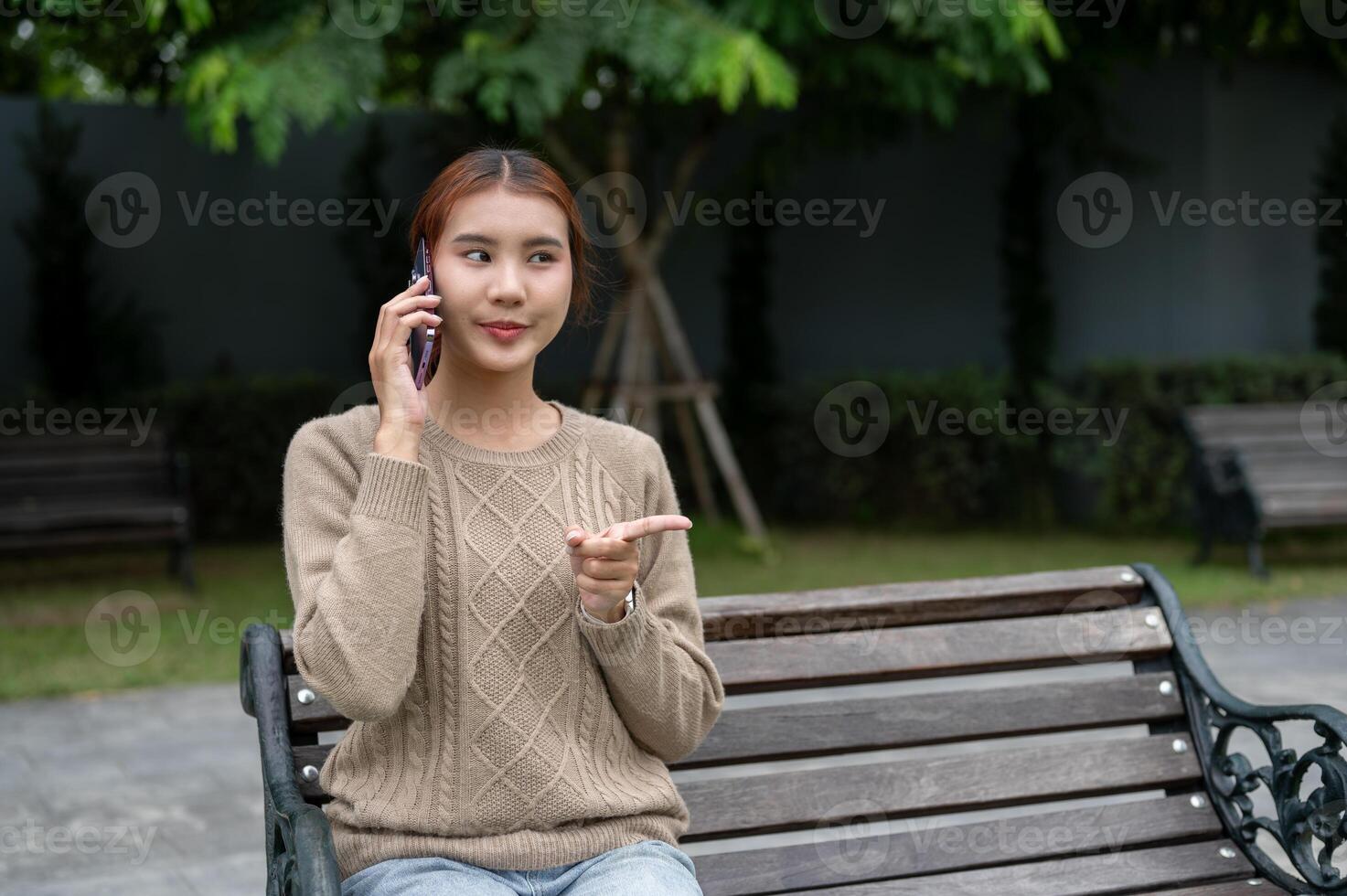 A beautiful Asian woman is talking on the phone with someone while resting on a bench in a park. photo