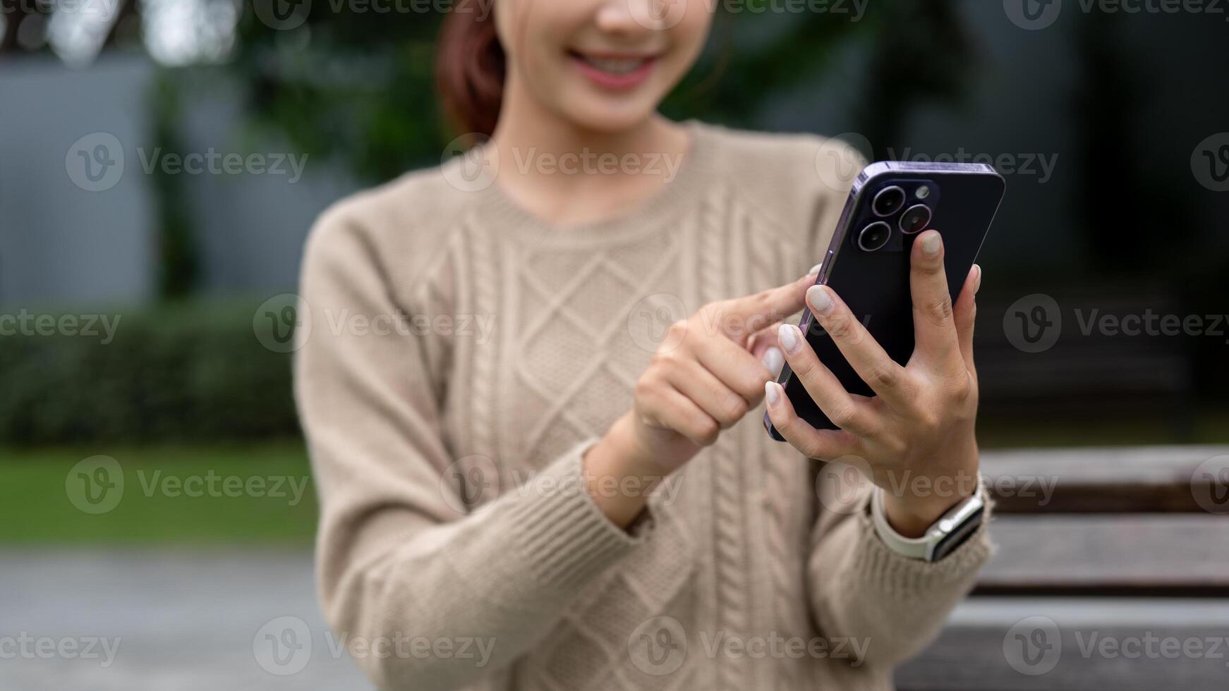 A close-up image of a happy Asian woman is using her smartphone on a bench in a garden. photo