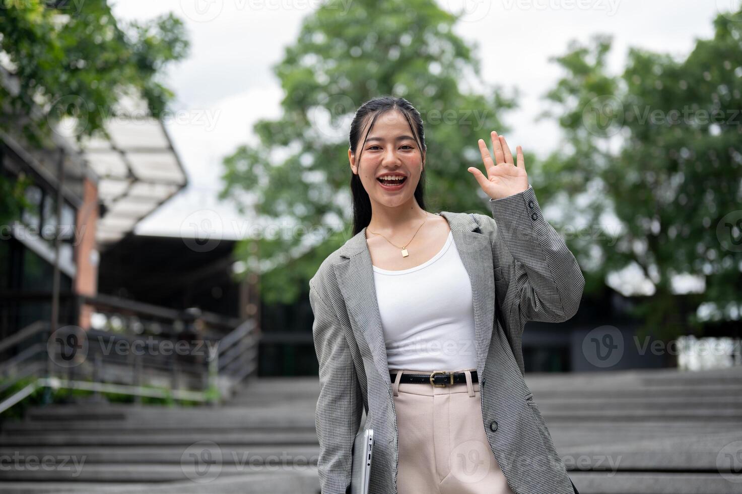 Cheerful Asian businesswoman is waving her hand and smiling at the camera while standing in the city photo