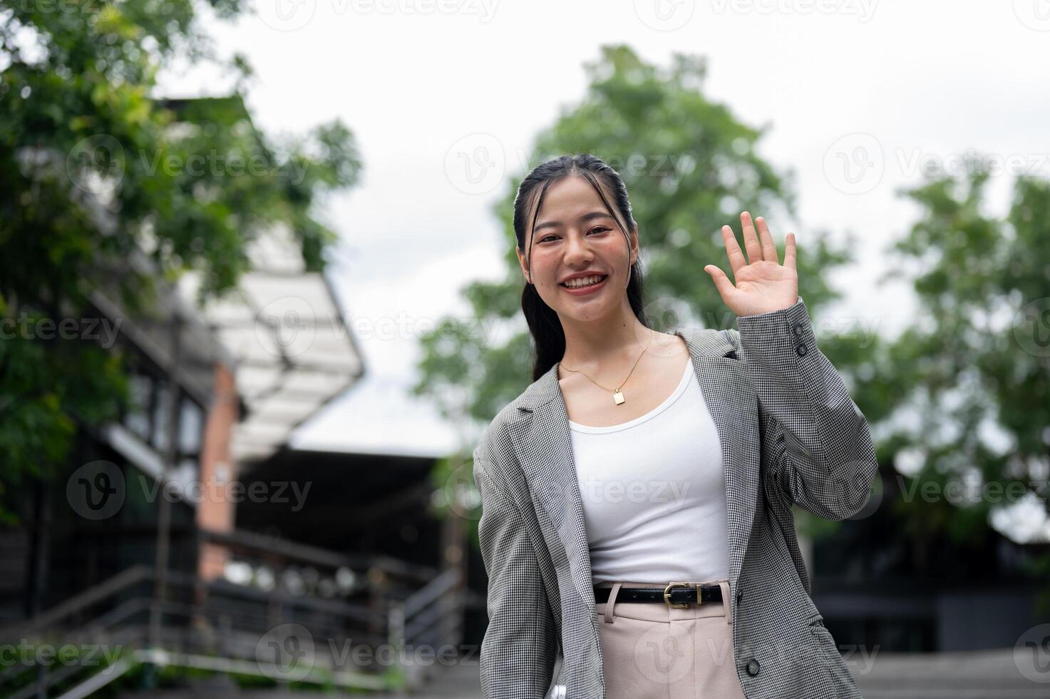 Cheerful Asian businesswoman is waving her hand and smiling at the camera while standing in the city photo