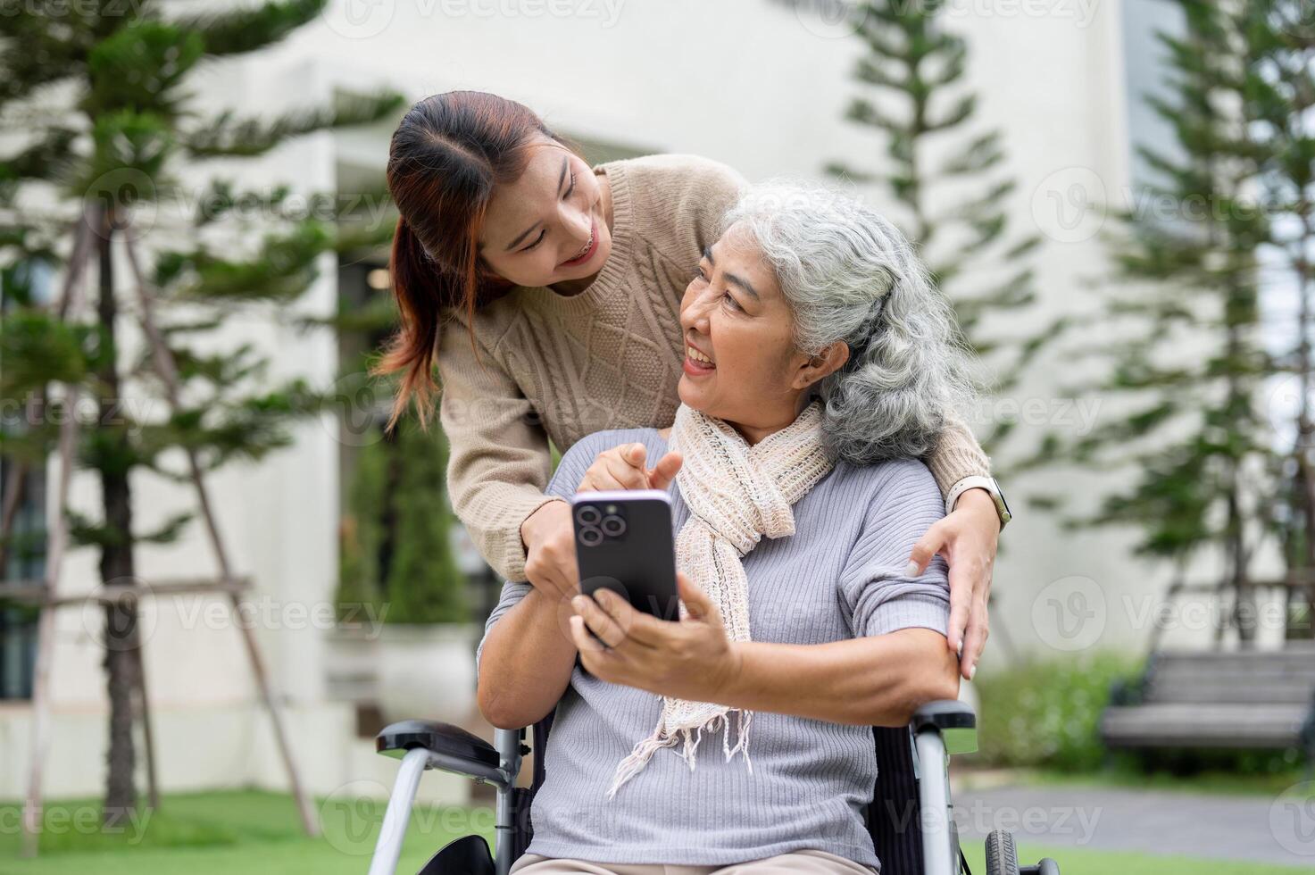 A happy Asian grandma in a wheelchair is showing her phone to her granddaughter. photo