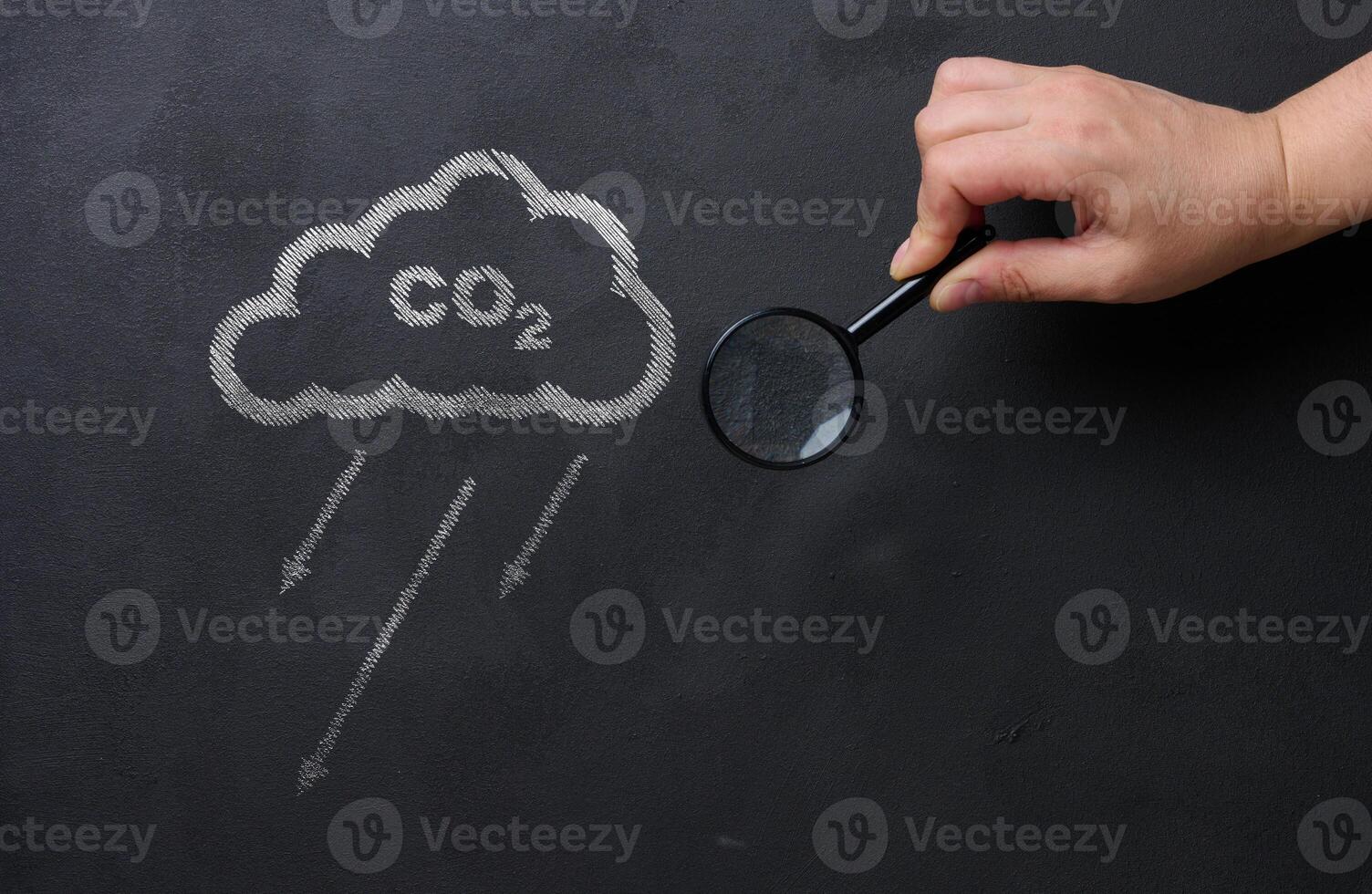 Female hand holds a magnifying glass against a black chalkboard background and a cloud, concept of reducing CO2 emissions photo