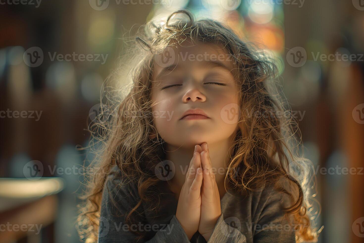 A little girl with curly hair, wearing gray and praying in the church with hands clasped together photo