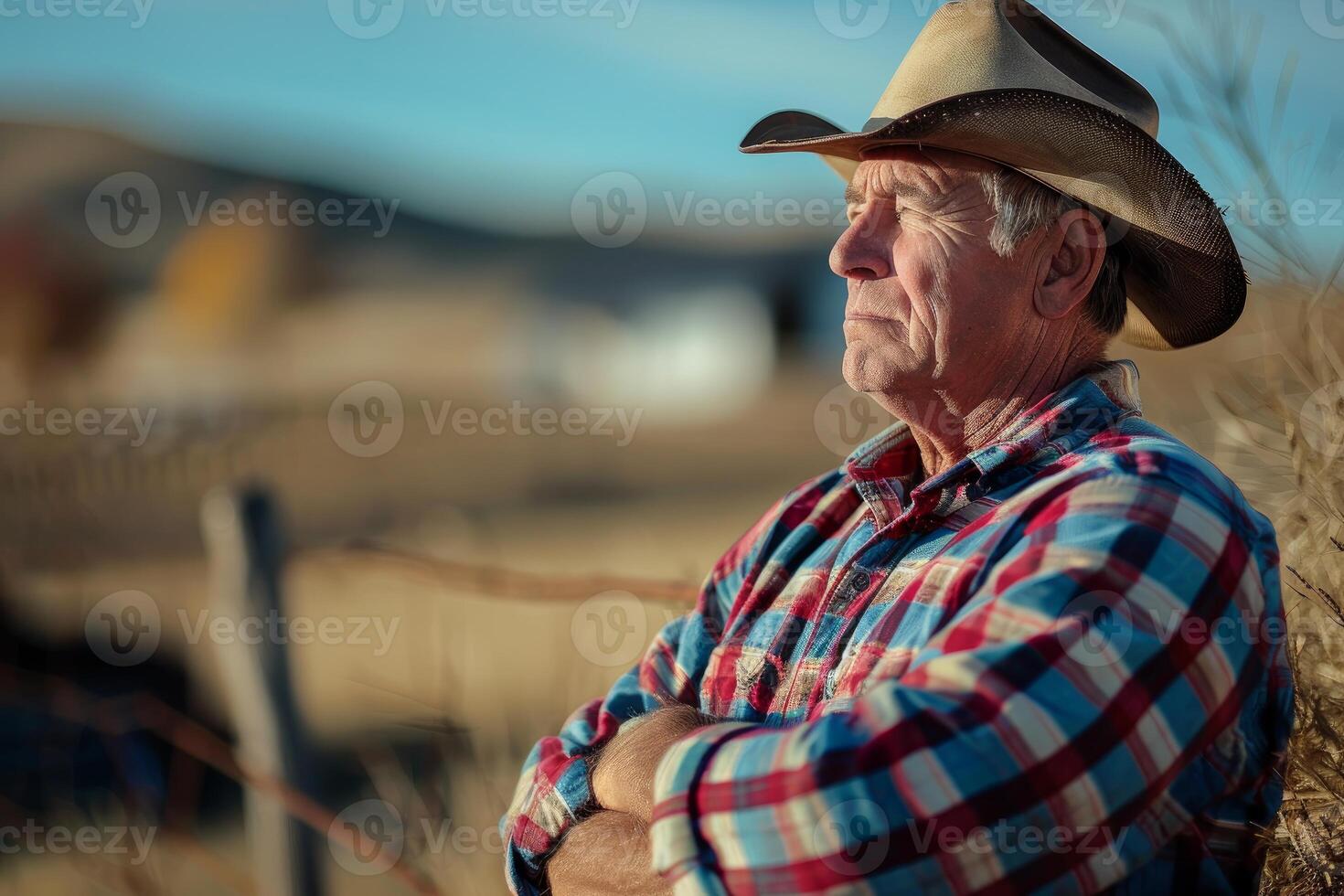 Elder cowboy in a hat stands pensively with crossed arms against a rural background photo