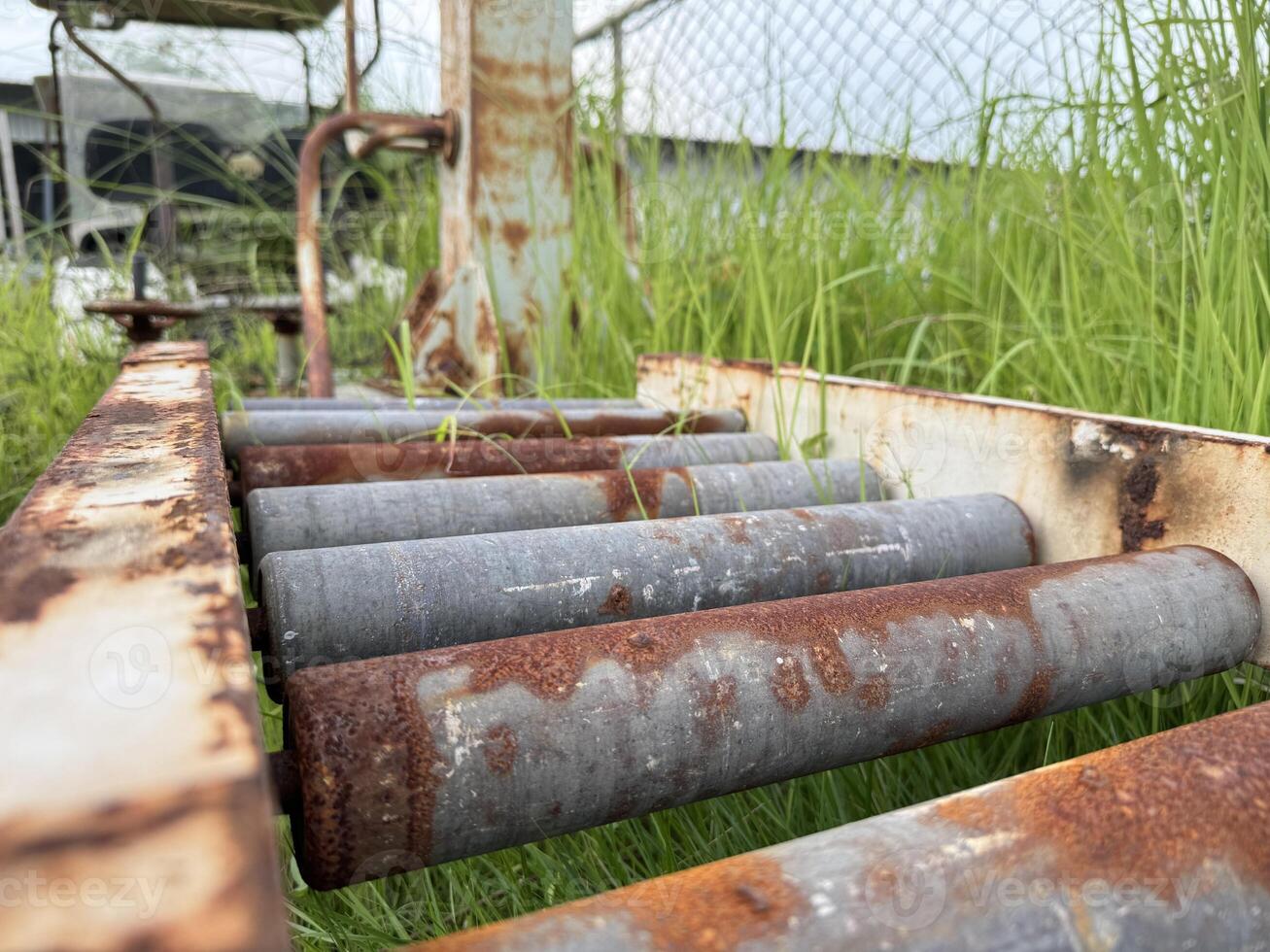 A close-up view of an aged, rusted roller conveyor, partially obscured by lush green grass, evokes a sense of industrial history and the passage of time. photo