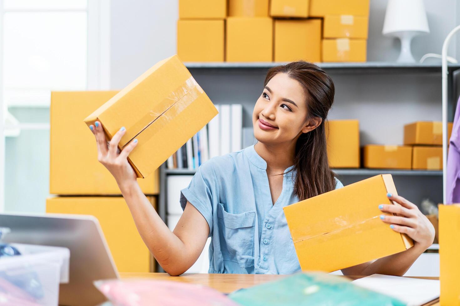 Entrepreneurs Small Business SME freelance, young Asian woman holding parcel box and sitting on chair, she is happy smile work at home, online marketing packing box delivery, online sales photo