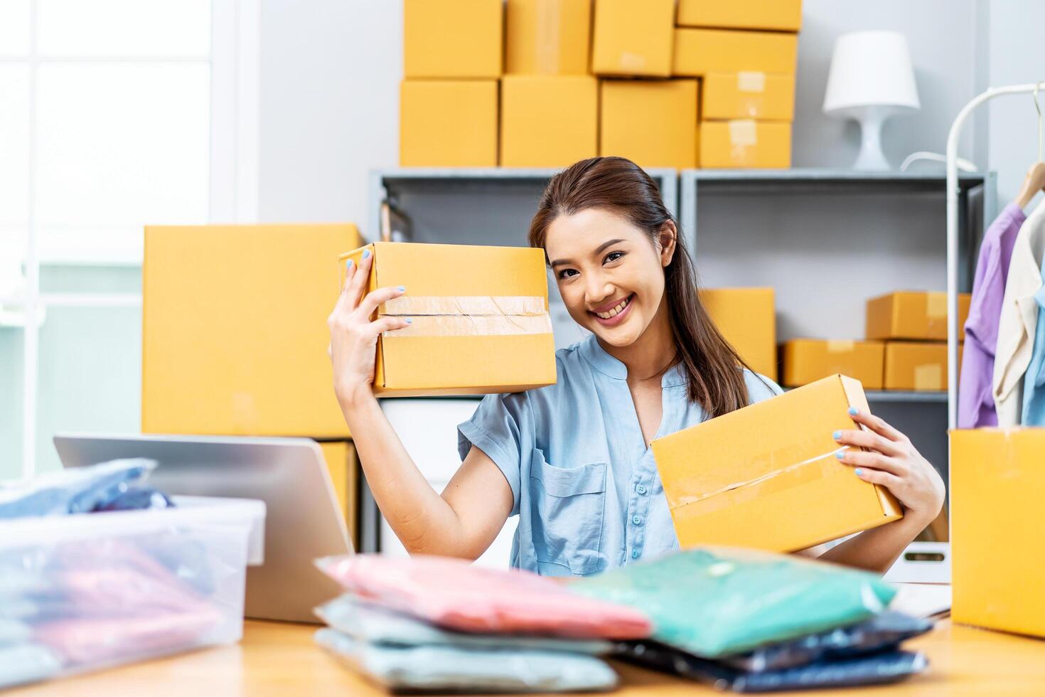 Entrepreneurs Small Business SME freelance, young Asian woman holding parcel box and sitting on chair, she is happy smile work at home, online marketing packing box delivery, online sales photo