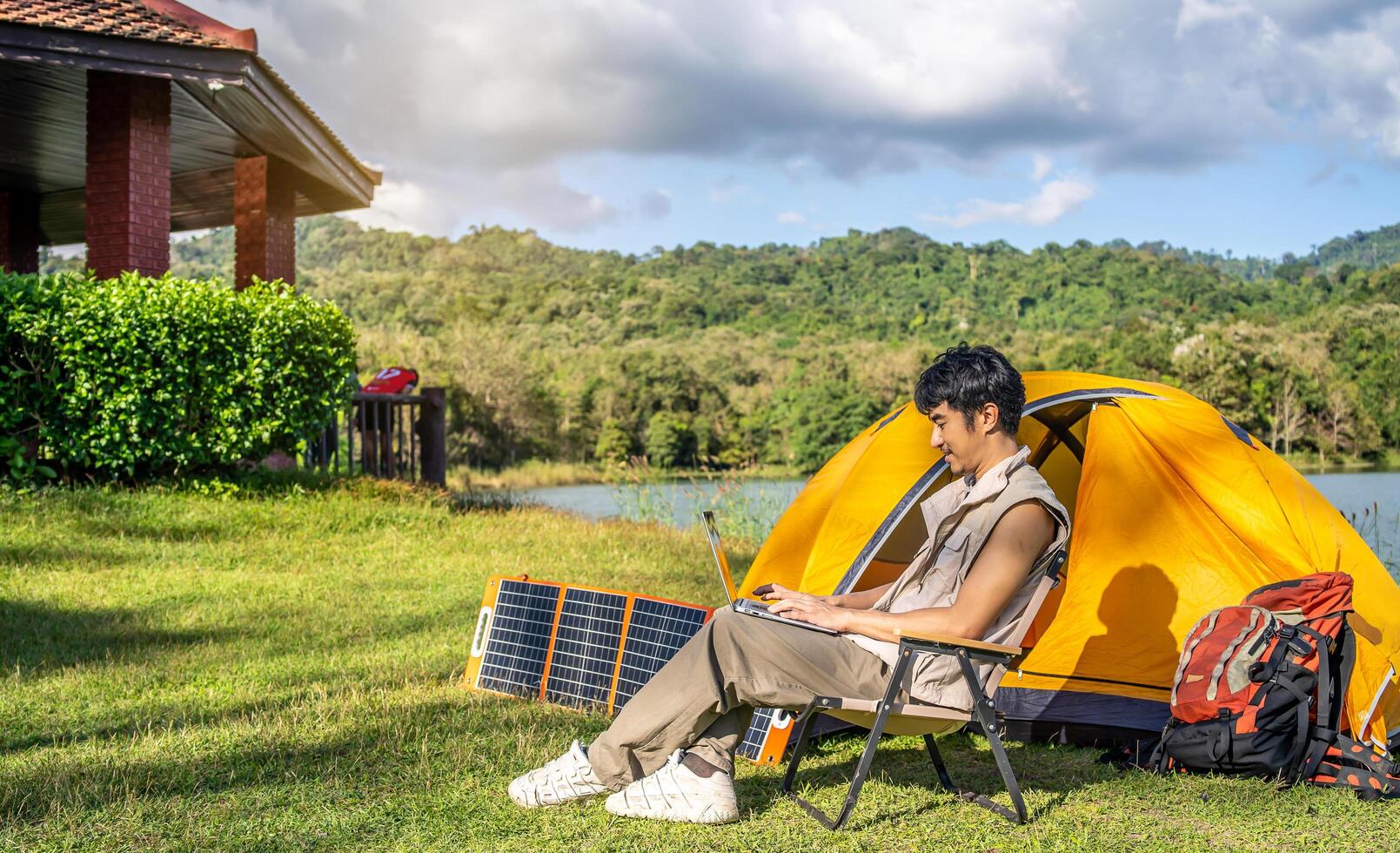 Young Asian traveling man relax and enjoy outdoor lifestyle camping in forest mountain on summer holiday. Attractive male relaxing in chair while working on laptop during camping day in nature photo