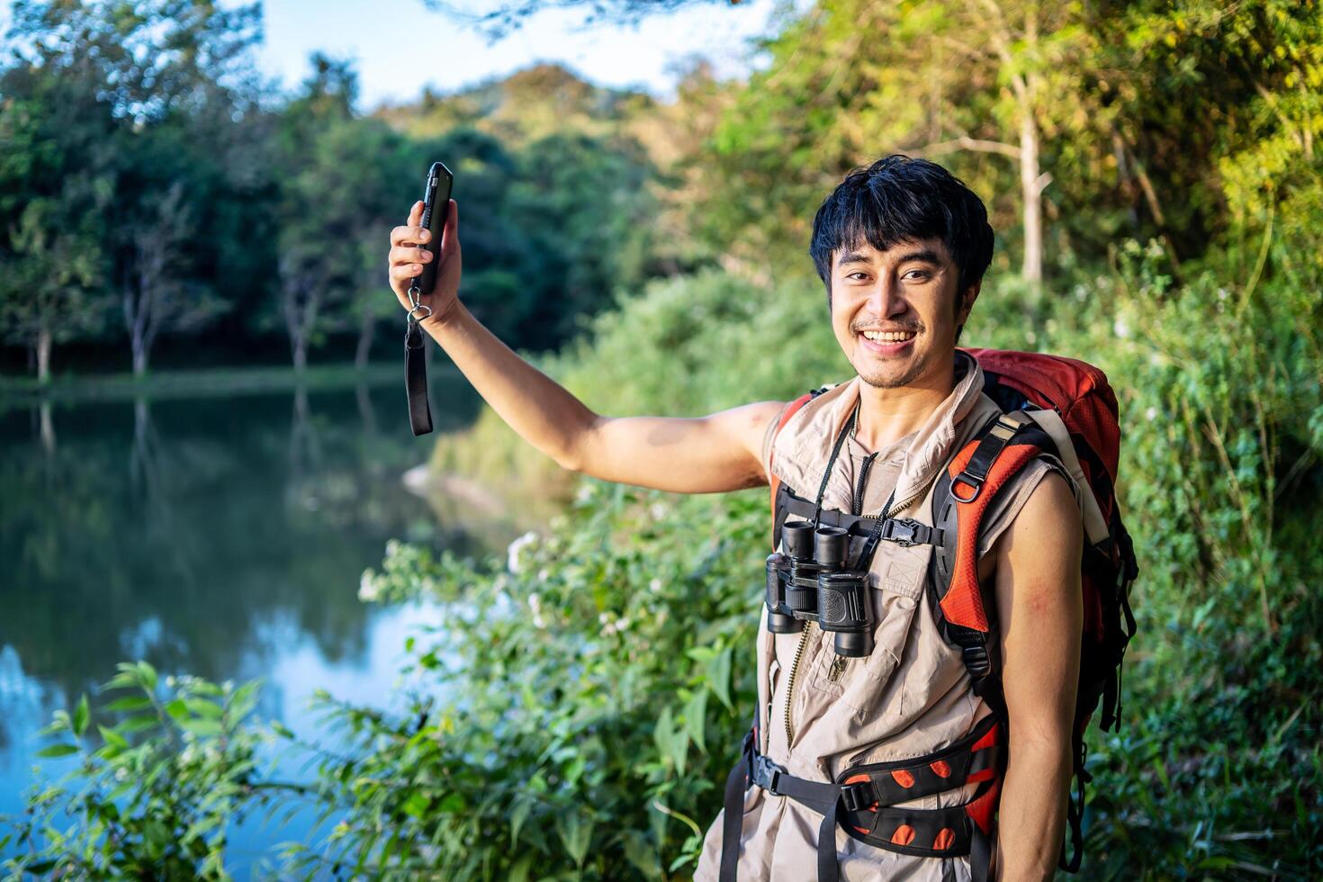 Young asian attractive male tourists takes a photo of a beautiful autumn nature with a view of a lake and a park with his phone and selfie. Walking in the park alone, admiring the beauty nature