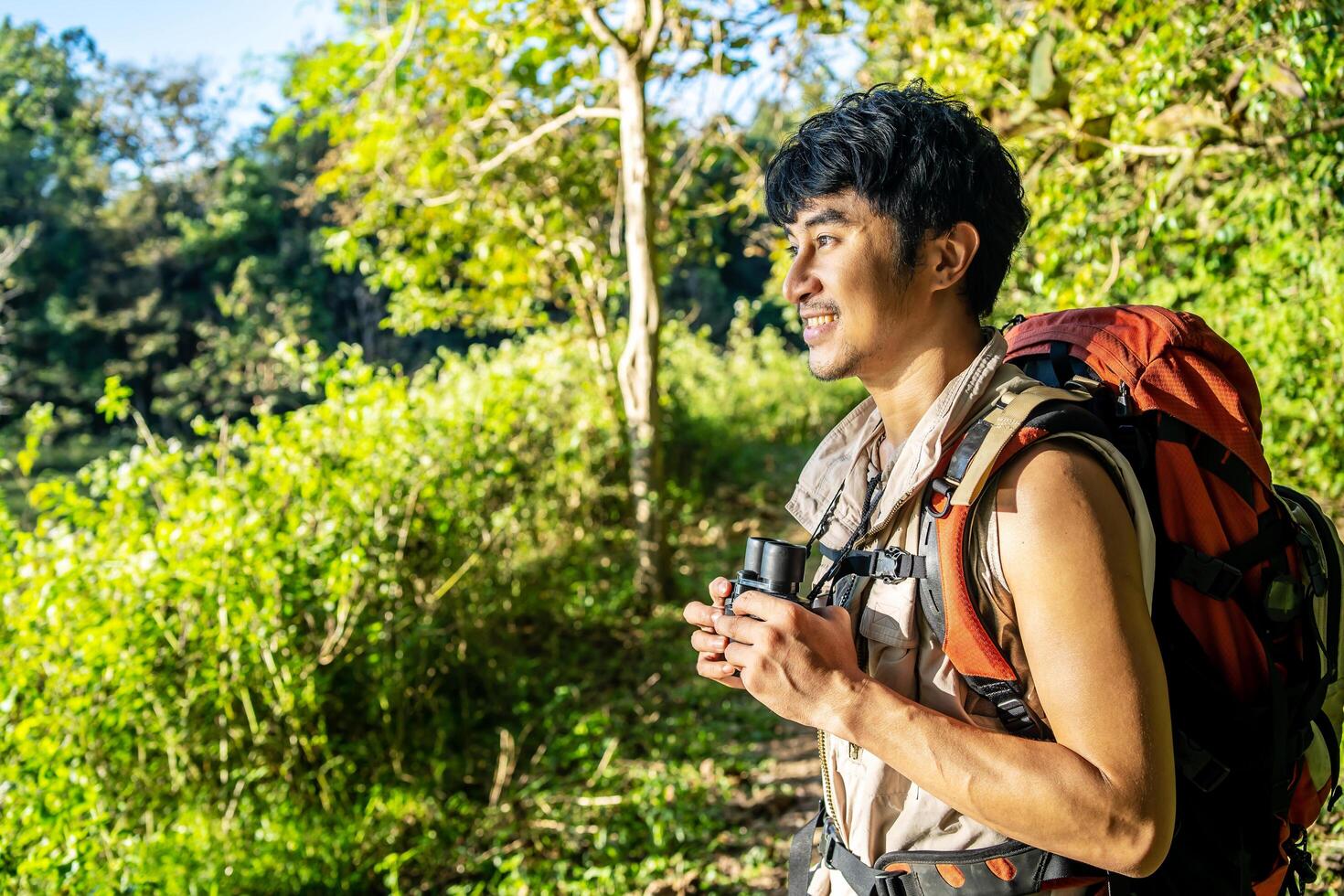 Young travel man relax and enjoy in the Woods, holds binoculars during a camping trip against blue vivid sky. photo