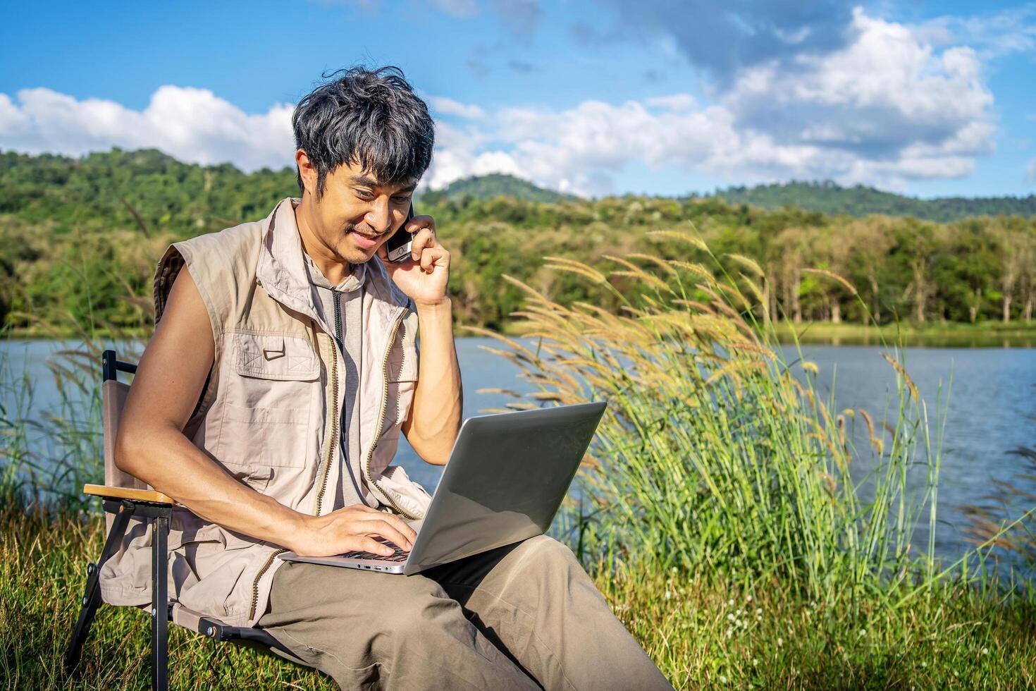 Young happy asian travel male sitting on chair in park, talking on mobile phone and tapping on laptop. Man using computer and having call on cellphone outdoor on sunny fall day. Freelancer working photo