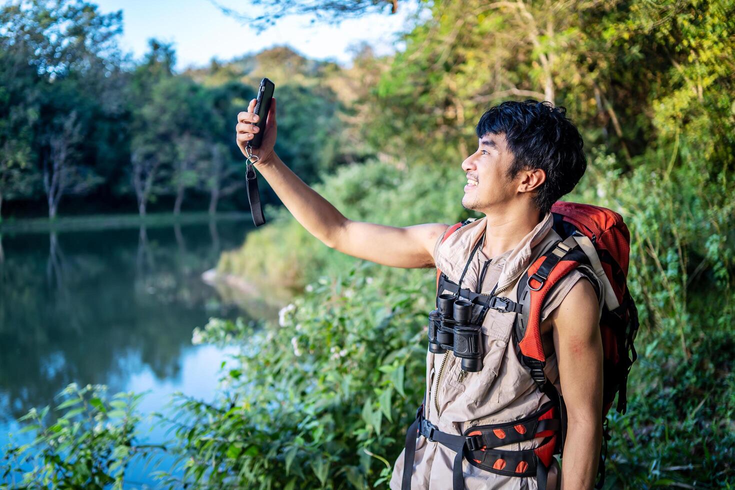 Young asian attractive male tourists takes a photo of a beautiful autumn nature with a view of a lake and a park with his phone and selfie. Walking in the park alone, admiring the beauty nature