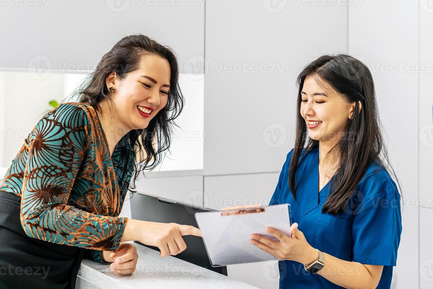 Asian woman comes to reception desk in clinic lobby area and makes appointment with doctor. Female administrator talks with patient at information counter. Medical staff work in medical center photo