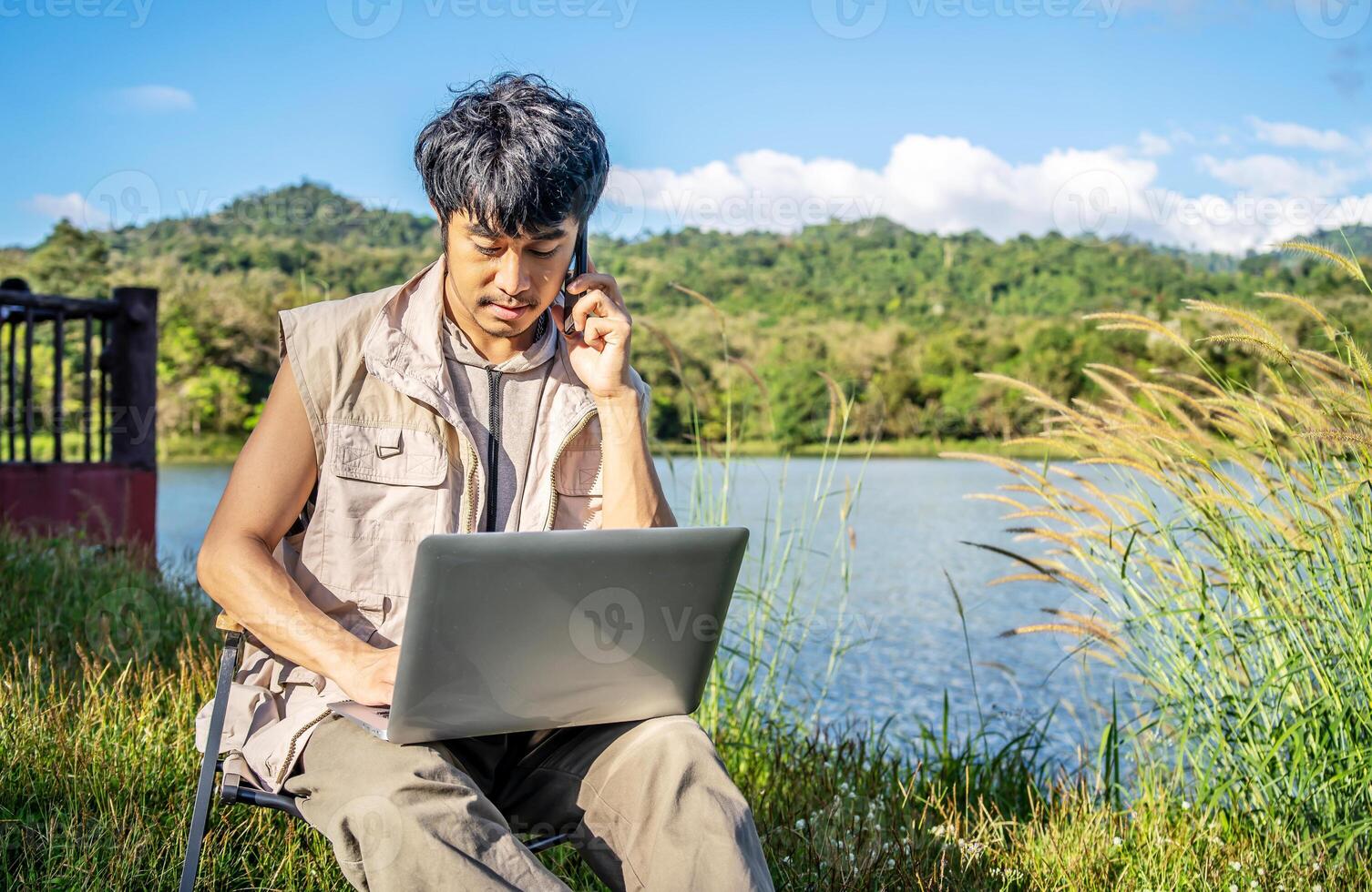 Young happy asian travel male sitting on chair in park, talking on mobile phone and tapping on laptop. Man using computer and having call on cellphone outdoor on sunny fall day. Freelancer working photo
