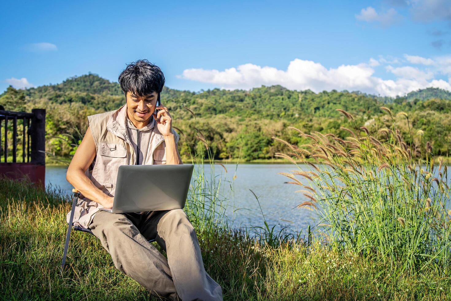Young happy asian travel male sitting on chair in park, talking on mobile phone and tapping on laptop. Man using computer and having call on cellphone outdoor on sunny fall day. Freelancer working photo