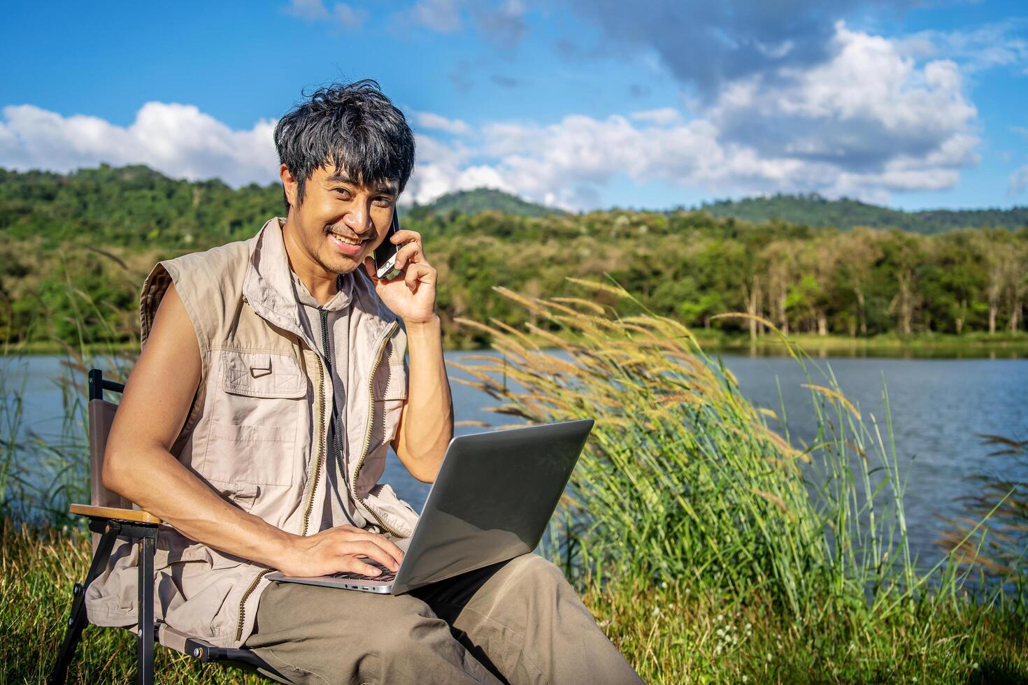 Young happy asian travel male sitting on chair in park, talking on mobile phone and tapping on laptop. Man using computer and having call on cellphone outdoor on sunny fall day. Freelancer working photo