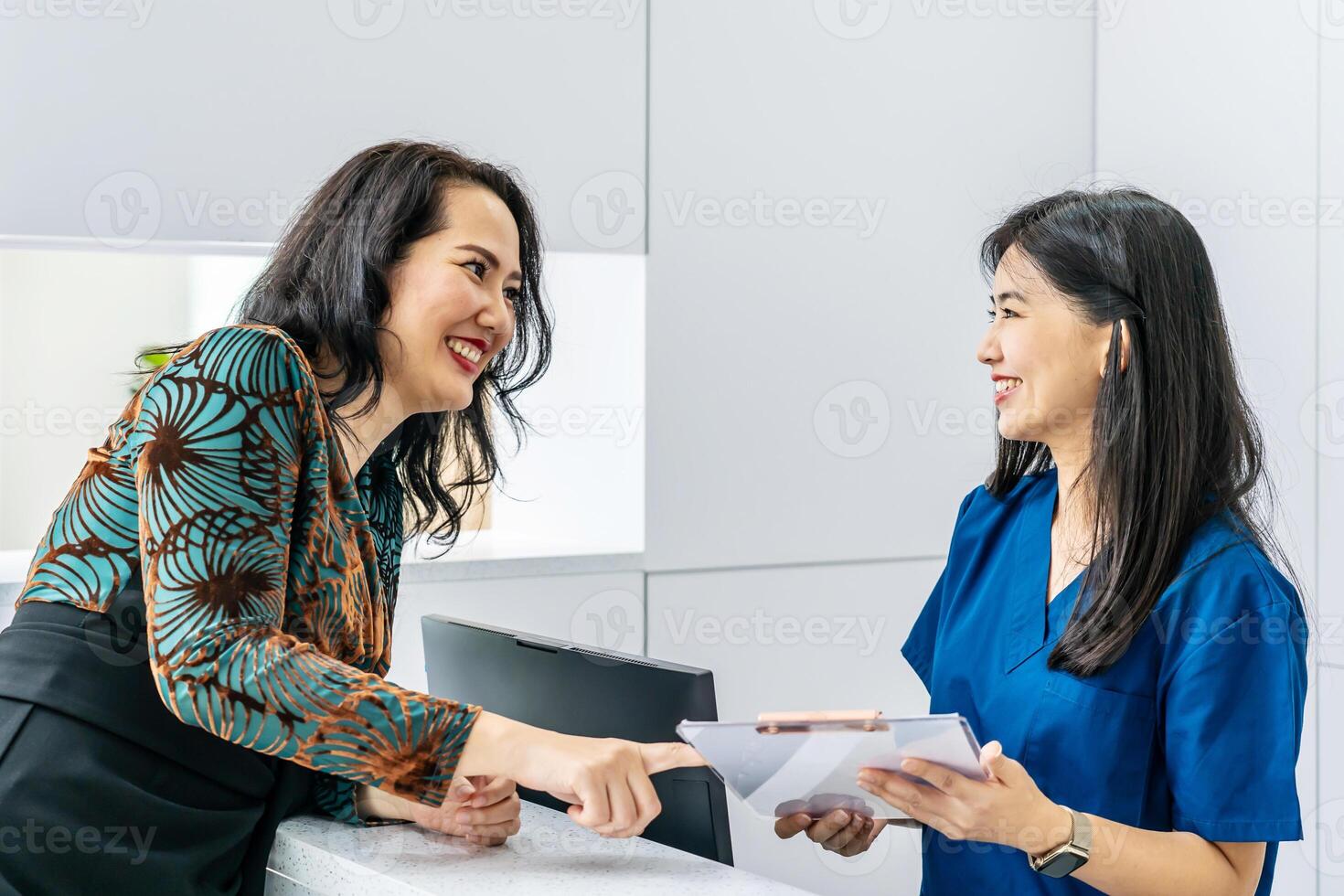Asian woman comes to reception desk in clinic lobby area and makes appointment with doctor. Female administrator talks with patient at information counter. Medical staff work in medical center photo