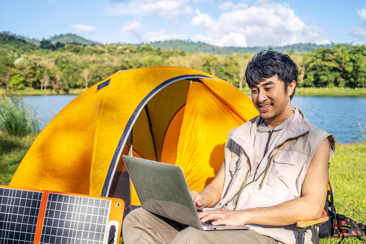 Young Asian traveling man relax and enjoy outdoor lifestyle camping in forest mountain on summer holiday. Attractive male relaxing in chair while working on laptop during camping day in nature photo