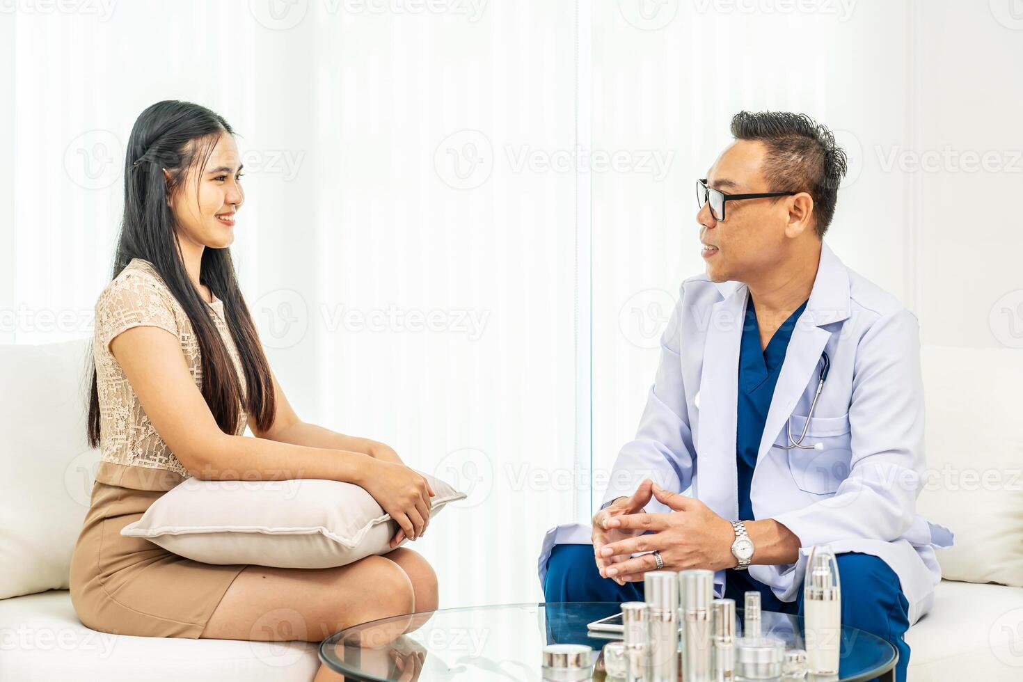Consultant man with wearing medical gown showing creams and lotions from desk to young asian female. Dermatologist giving advice on proper skin care product. Concept of beauty, Experts, Cosmetics. photo
