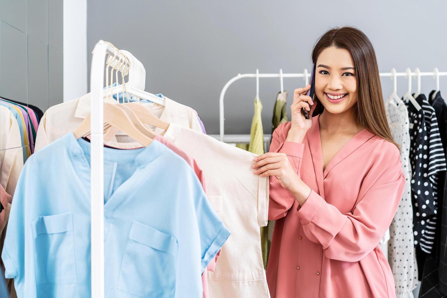 Beautiful asian woman online seller confirming orders from customer on the phone. E-commerce business owner looking and choose clothes in the clothes rail with talking on phone in store warehouse. photo