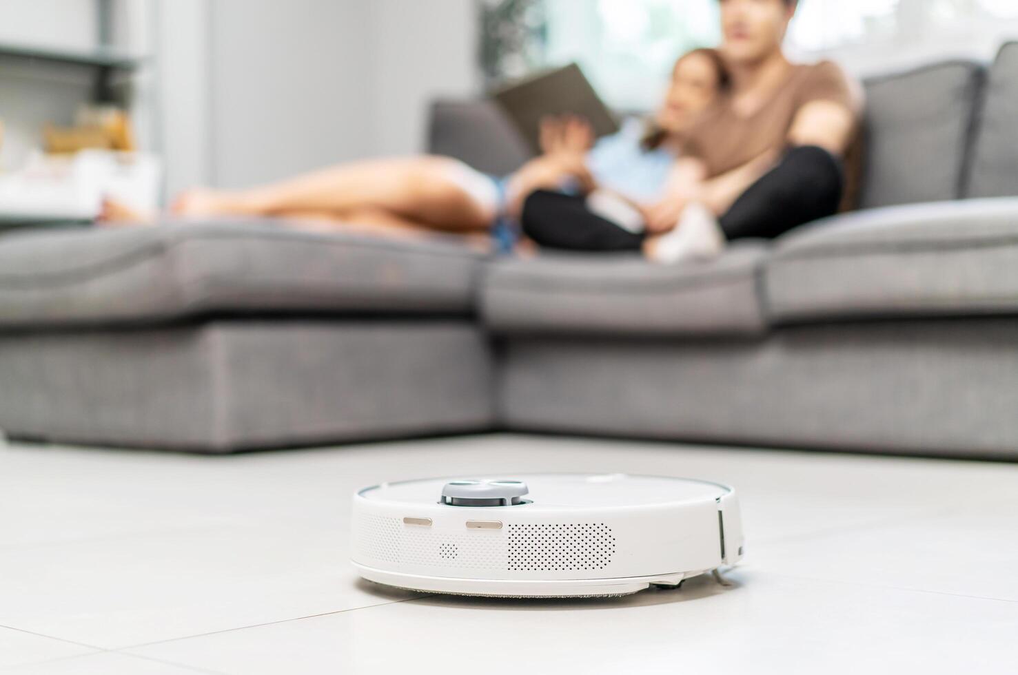 Young couple resting on the couch while robotic vacuum cleaner doing its work at home. Household robots, Smart home concept photo