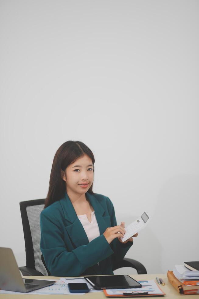 Asian businesswoman in red suit calculating company sales with calculator, laptop, and tablet on table Interior of businesswoman office at the modern workstation. photo