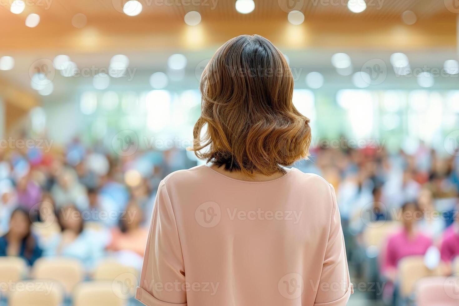 Rear view of a woman speaking to a diverse audience in a brightly lit conference hall, focusing on professional communication. photo
