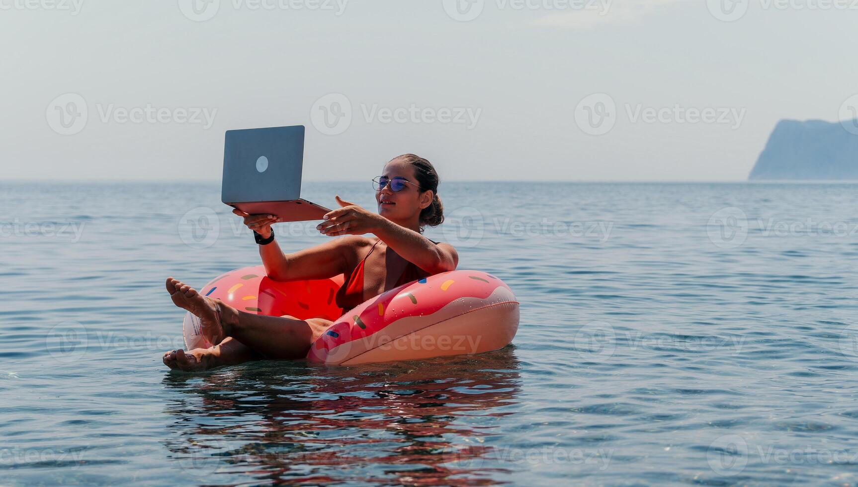 Woman freelancer works on laptop swimming in sea on pink inflatable ring. Happy tourist floating on inflatable donut and working on laptop computer in calm ocean. Freelance, remote working anywhere photo