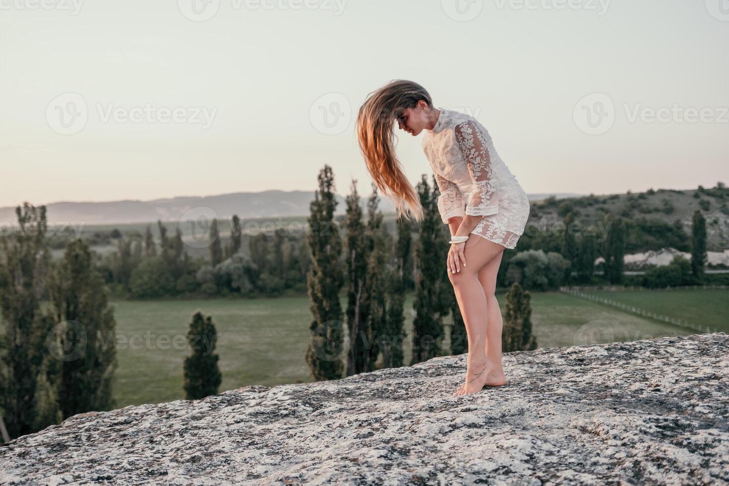 Happy woman in white boho dress on sunset in mountains. Romantic woman with long hair standing with her back on the sunset in nature in summer with open hands. Silhouette. Nature. Sunset. photo