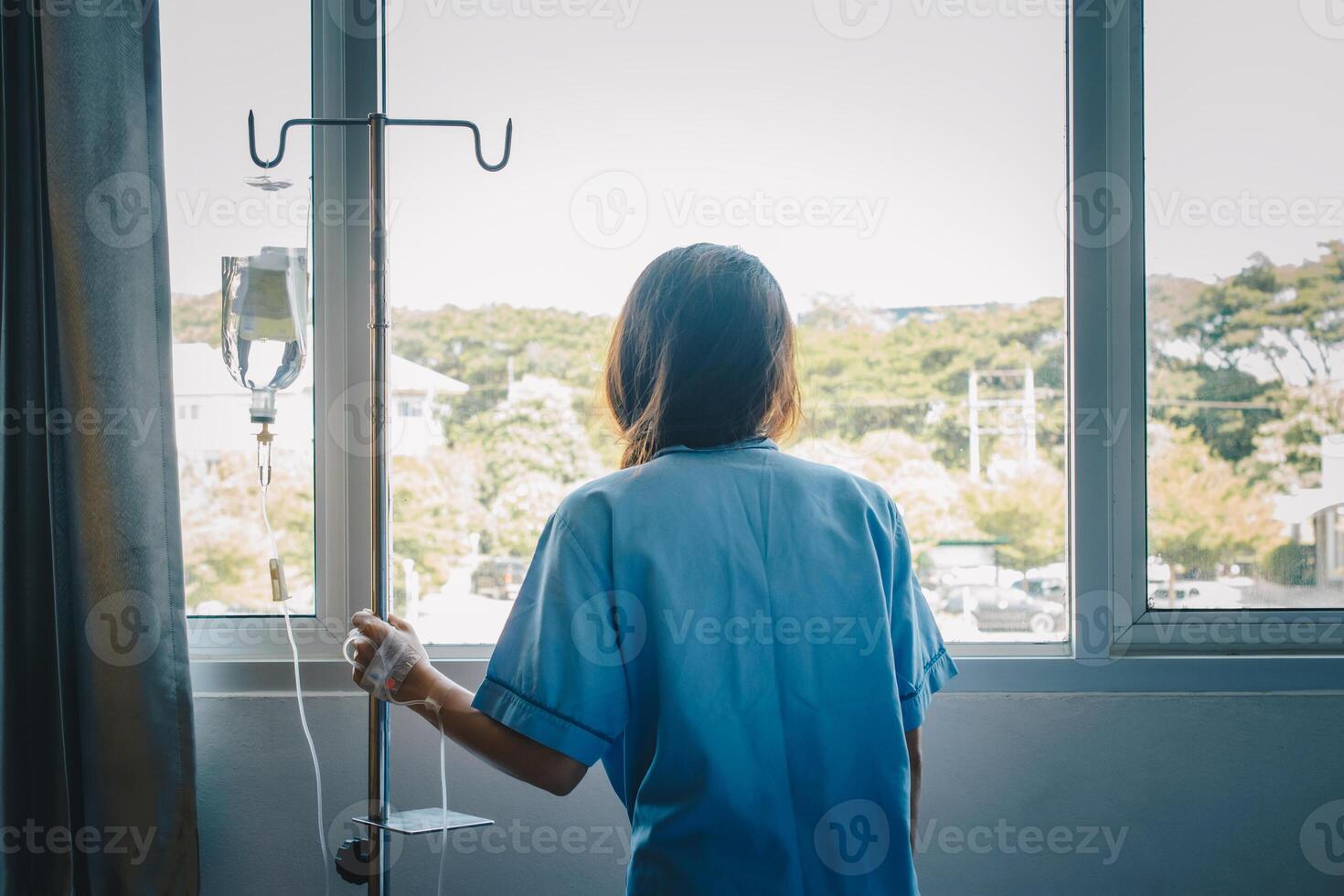 Back view of sickness patient woman standing nearly window in hospital ward, looking to outside and hope her health will be better. photo